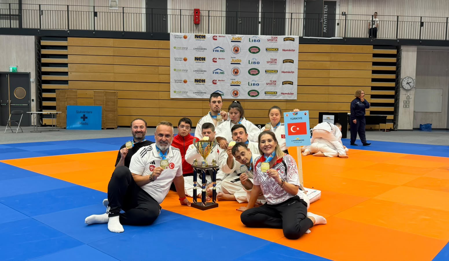Milli takım sporcuları ve teknik ekip, tatami üzerinde kupa ve madalyalarla birlikte poz veriyor.