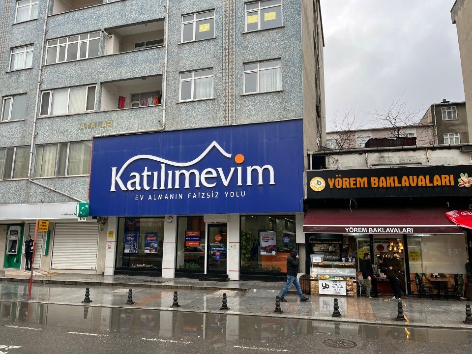 Yağmurlu bir cadde üzerinde Katılımevim tabelalı şube ve çevredeki dükkânlar.