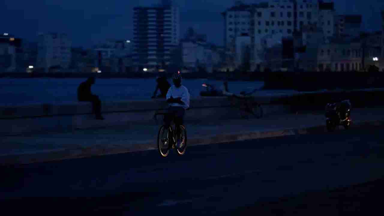 A person riding a bicycle on a dark coastline and a dimly lit city silhouette in the background.