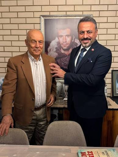 Bora Gezmiş ve Metin Demirbilek, arkalarında Deniz Gezmiş fotoğrafı bulunan bir odada yan yana duruyor.