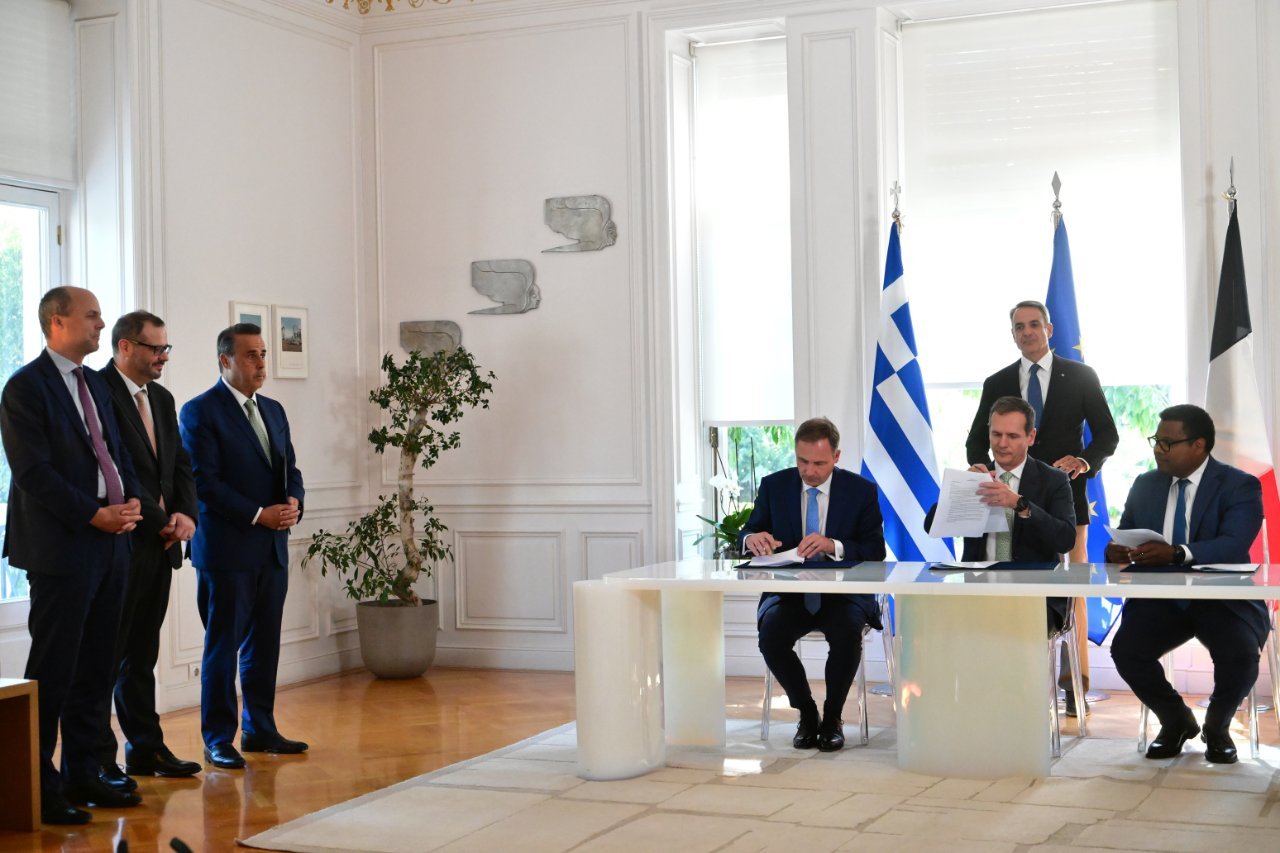 Funzionari firmano documenti in una sala con bandiere greche ed europee.