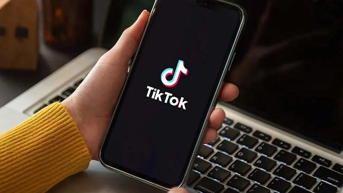Bir kişinin elinde TikTok logosu görünen telefon ve arka planda dizüstü bilgisayar.