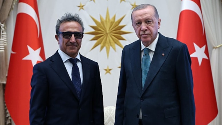 Hamdi Ulukaya ve Cumhurbaşkanı Recep Tayyip Erdoğan, Türk bayraklarının bulunduğu bir salonda yan yana duruyor.
