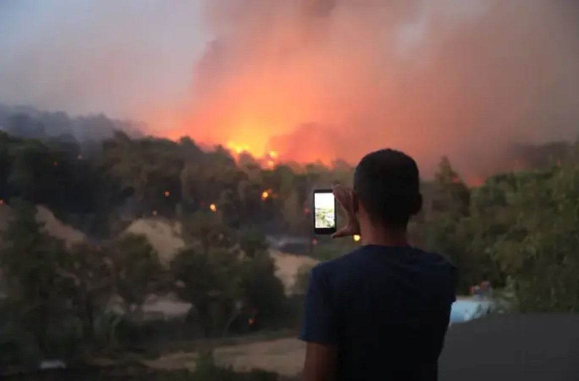 A moment where a person is filming the distant rising flames and smoke with a mobile phone.
